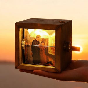 May include: A wooden photo cube with a picture of a couple kissing under an umbrella. The cube has a small crank on the side and is held in a hand against a sunset backdrop. The cube is illuminated from within.