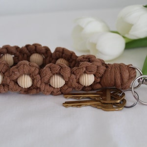 May include: A brown macrame keychain with a floral design, featuring wooden beads at the center of each flower. The keychain is attached to a silver metal ring and clasp, with two gold keys. White tulips are in the background.