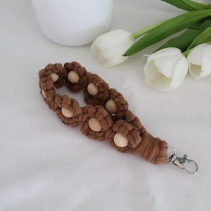 Pu&ograve; includere: Un cinturino da polso in macram&egrave; marrone con inserti di perline di legno e chiusura color argento. Il cinturino &egrave; decorato con nodi a forma di fiore. Tulipani bianchi e un vaso bianco sullo sfondo.