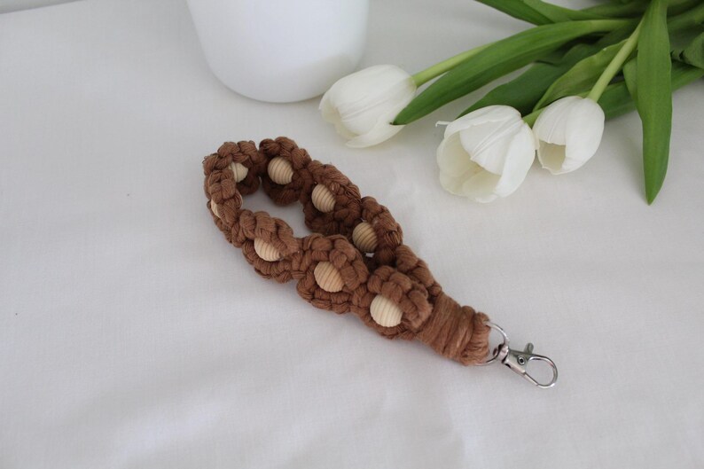 Pu&ograve; includere: Un cinturino da polso in macram&egrave; marrone con inserti di perline di legno e chiusura color argento. Il cinturino &egrave; decorato con nodi a forma di fiore. Tulipani bianchi e un vaso bianco sullo sfondo.