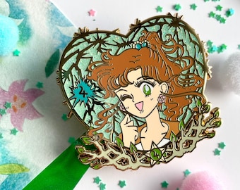 May include: A gold-colored enamel pin in the shape of a heart, featuring a cartoon character with brown hair and green eyes. The pin has a green and gold background with the number "24" and is surrounded by a leafy design.