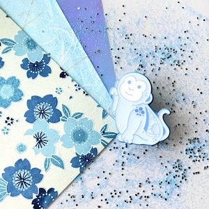 May include: A light blue enamel pin of a monkey with a white face and white eyes. The monkey is sitting on a white background with blue glitter.