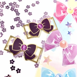 Puede incluir: Dos pines de esmalte de color dorado en forma de lazos con un centro morado y un círculo rosa con la letra "t" y el número "2" en su interior. Los pines están sobre un fondo blanco con purpurina morada.