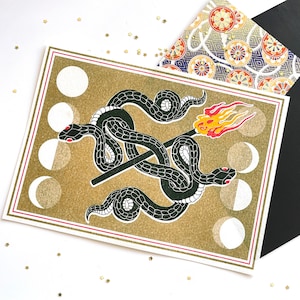 May include: A rectangular greeting card featuring a gold glitter background with two black snakes intertwined around a lit matchstick. The design includes a series of white moon phases along the left and right borders and a red and white frame.
