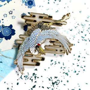 以下が含まれることがあります： 雲の中を飛ぶスタイリッシュな鶴をデザインした、金色と青色のエナメルピン。鶴は青と白の配色で、黒のアクセントが付けられています。雲はスタイリッシュで、金色の仕上げになっています。