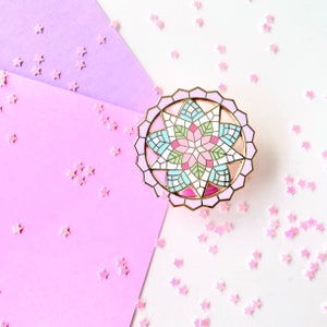 Puede incluir: Un pin redondo de esmalte con un diseño de vidriera. El pin presenta un borde dorado y un interior colorido con formas geométricas rosas, azules, verdes y blancas. El pin está sobre un fondo de papel rosa y blanco con confeti rosa en forma de estrella esparcido.