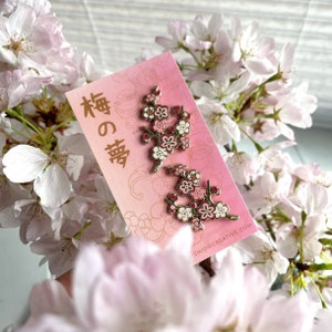 Puede incluir: Dos pines de esmalte dorado con flores de cerezo rosas y blancas. Los pines están en una tarjeta rosa y blanca con texto japonés que dice "桜の詩" que se traduce como "Poema de la flor de cerezo".