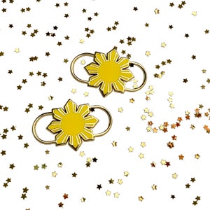 May include: Two gold-colored pins featuring a yellow sunburst design with white accents. The pins have a gold-colored ring attached. The background is white with scattered gold star-shaped confetti.