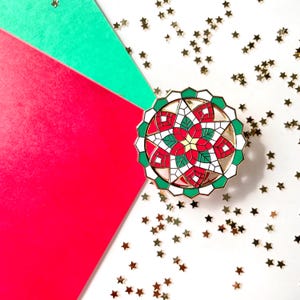 Puede incluir: Un pin redondo de esmalte con un diseño de vidriera con colores rojo, verde y blanco. El pin tiene un borde dorado y está colocado sobre un fondo de papel rojo, verde y blanco con confeti de estrellas doradas.