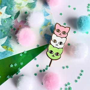 Op de afbeelding: Een gouden emaille pin met drie gestapelde kattengezichten op een stok. De katten zijn groen, wit en roze, en lijken op een Japanse dango.