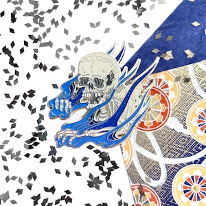 Op de afbeelding: Een zilveren emaille pin met een skelet met een schedel die uit blauwe vlammen komt. De vlammen hebben een glinsterende textuur.