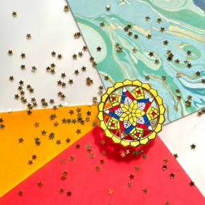 Puede incluir: Un pin de esmalte amarillo, rojo, azul y blanco con un diseño de estrella. El pin está sobre un fondo blanco, naranja y azul con purpurina dorada.