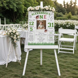 May include: A white wooden easel sign with floral accents, displaying a child's first birthday stats. The sign includes a photo, the name "Alexander's", and details like 31 inches, 12 months, and 5 teeth. The sign is decorated with green grass.