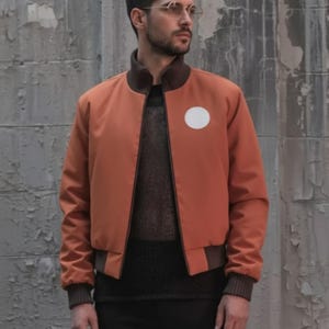 May include: A burnt orange bomber jacket with a white circular patch on the chest. The jacket has brown ribbed cuffs, collar, and hem. The person is wearing a dark sweater and black pants.