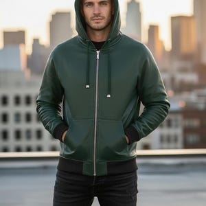 May include: A dark green hooded jacket with a zipper closure, front pockets, and black ribbed cuffs and hem. The jacket is worn with black jeans. The background features a cityscape.