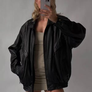 May include: Black leather jacket with a classic design. The jacket features a collared neckline, zipper closure, and pockets. The jacket is worn over a beige dress. The overall style is casual and fashionable.