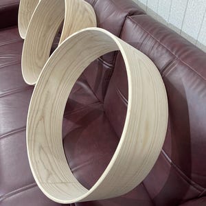 Puede incluir: Tres aros de tambor de madera sin terminar, que muestran la veta natural de la madera de color claro. Los aros están colocados sobre un sofá de cuero burdeos, destacando la forma circular y la textura de la madera.