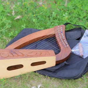 Può includere: Un'arpa lira in legno con corde, una base in legno separata e una custodia nera. L'arpa è realizzata in legno marrone scuro con piroli argentati. Un sacchetto di plastica trasparente con accessori è all'interno della custodia.