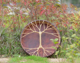 Può includere: Un tamburo rotondo marrone con un disegno dell'albero della vita al centro. Il tamburo è fatto di pelle e legno, con un bordo intrecciato. Lo sfondo presenta erba verde e fogliame colorato.