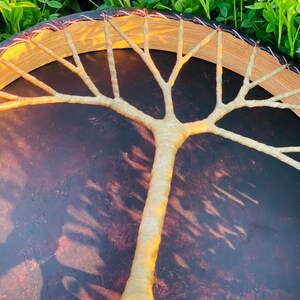 May include: A handcrafted drum with a tree design. The drum features a dark brown surface with a light brown tree design in the center. The drum's frame is a light brown color with a dark brown trim.