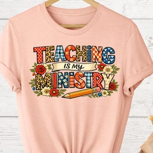 May include: A light peach-colored t-shirt with the phrase "TEACHING IS MY MINISTRY" in a colorful, patterned design. The design includes floral accents and a pencil illustration. The shirt is displayed on a wooden hanger.