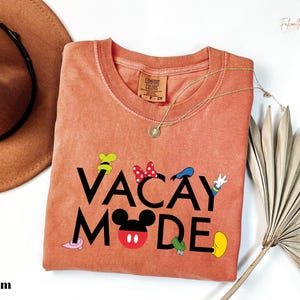 a pink t-shirt with the text "VACAY MODE" printed on it, along with various Disney character icons. The t-shirt is laid out on a white surface, accompanied by a hat, a necklace, and a fan.