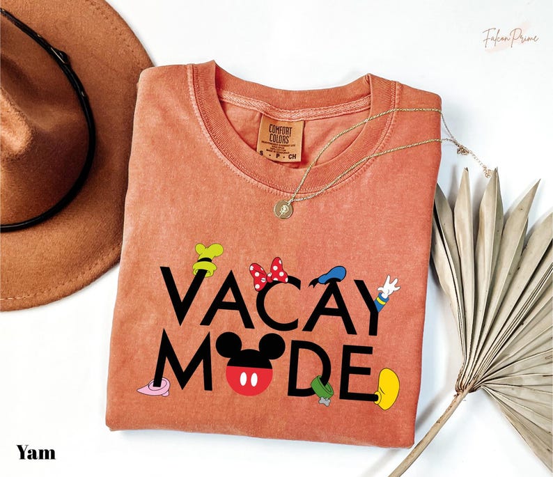 a pink t-shirt with the text "VACAY MODE" printed on it, along with various Disney character icons. The t-shirt is laid out on a white surface, accompanied by a hat, a necklace, and a fan.