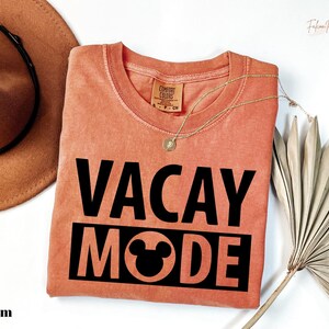 a pink t-shirt with the text "VACAY MODE" printed on it, along with a Mickey Mouse logo. The t-shirt is laid out on a white surface, and there is a hat and a fan nearby.