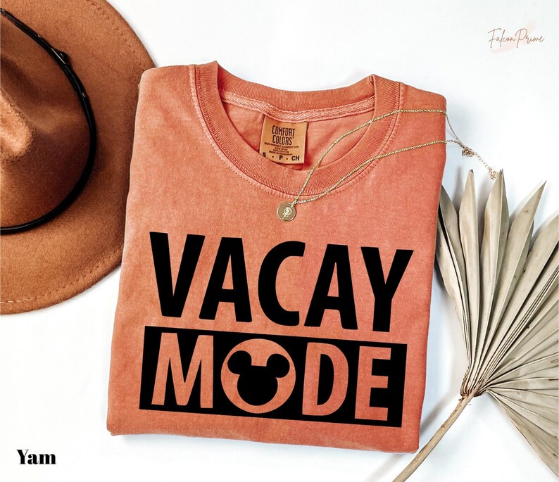 a pink t-shirt with the text "VACAY MODE" printed on it, along with a Mickey Mouse logo. The t-shirt is laid out on a white surface, and there is a hat and a fan nearby.