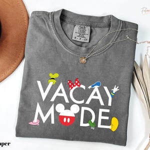 A gray t-shirt with the text "VACAY MODE" printed on it, along with various Disney character icons, is displayed on a white surface.