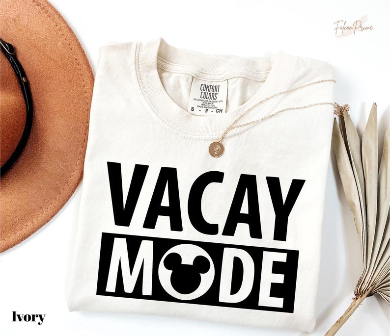 A white t-shirt with the text "VACAY MODE" printed on it, along with a small pendant necklace. The t-shirt is laid out on a white background, and there is a brown hat and a fan visible in the image.