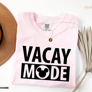A pink t-shirt with the text "VACAY MODE" printed on it, along with a Mickey Mouse design, is displayed on a white background. There is also a brown hat and a palm frond visible in the image.