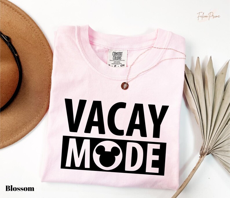 A pink t-shirt with the text "VACAY MODE" printed on it, along with a Mickey Mouse design, is displayed on a white background. There is also a brown hat and a palm frond visible in the image.