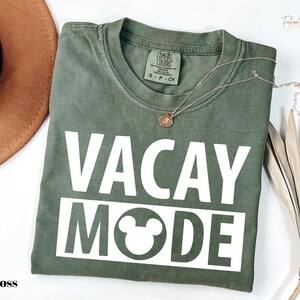 a green t-shirt with the text "VACAY MODE" printed on it, along with a Mickey Mouse symbol. The t-shirt is laid out on a white surface, and there is a brown hat and some dried leaves nearby.