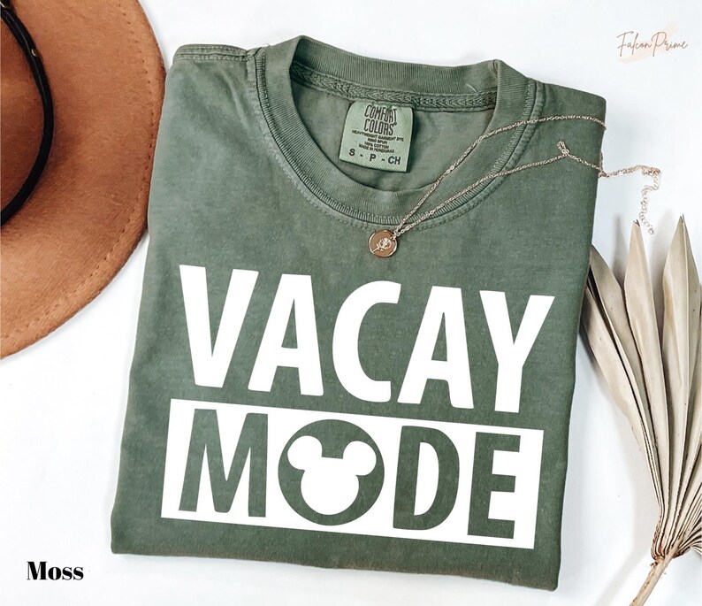 a green t-shirt with the text "VACAY MODE" printed on it, along with a Mickey Mouse symbol. The t-shirt is laid out on a white surface, and there is a brown hat and some dried leaves nearby.