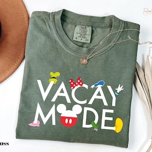 a green t-shirt with the text "VACAY MODE" printed on it, along with various Disney character illustrations. The t-shirt is laid out on a white surface, and there is a brown hat and some dried leaves nearby.
