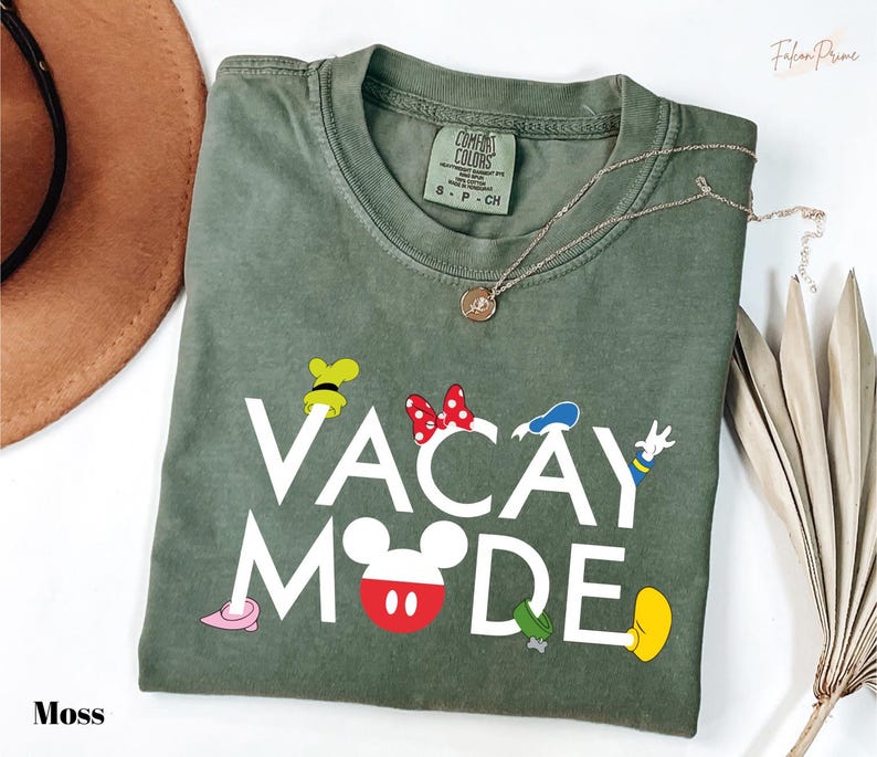 a green t-shirt with the text "VACAY MODE" printed on it, along with various Disney character illustrations. The t-shirt is laid out on a white surface, and there is a brown hat and some dried leaves nearby.