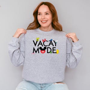Puede incluir: Una sudadera gris con el texto "VACAY MODE" en letras negras. El texto est&aacute; rodeado de personajes de dibujos animados coloridos, incluido Mickey Mouse.