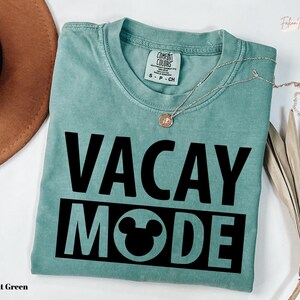 A light green t-shirt with the text "VACAY MODE" printed on it, along with a small pendant necklace. The t-shirt is folded and placed on a white surface, with a brown hat and some dried leaves nearby.