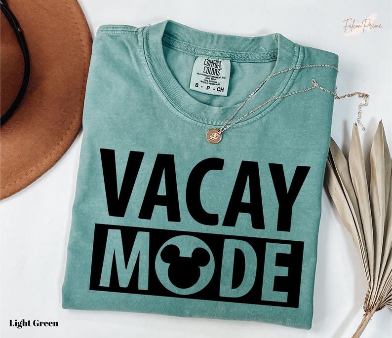 A light green t-shirt with the text "VACAY MODE" printed on it, along with a small pendant necklace. The t-shirt is folded and placed on a white surface, with a brown hat and some dried leaves nearby.