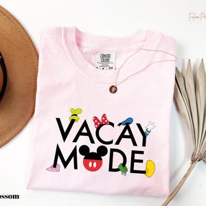 A pink t-shirt with the text "VACAY MODE" printed on it, along with various Disney character icons, is displayed on a white background.