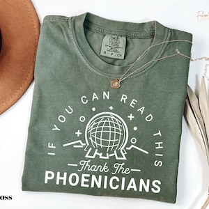 May include: Moss green t-shirt with the text "IF YOU CAN READ THIS - Thank The PHOENICIANS" in white. The shirt features a graphic of a globe and a Comfort Colors tag. A gold necklace is draped across the shirt.
