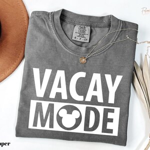 a gray t-shirt with the text "VACAY MODE" printed on it, along with a small pendant necklace. The t-shirt is laid out on a white surface, and there is a brown hat and some dried leaves nearby.