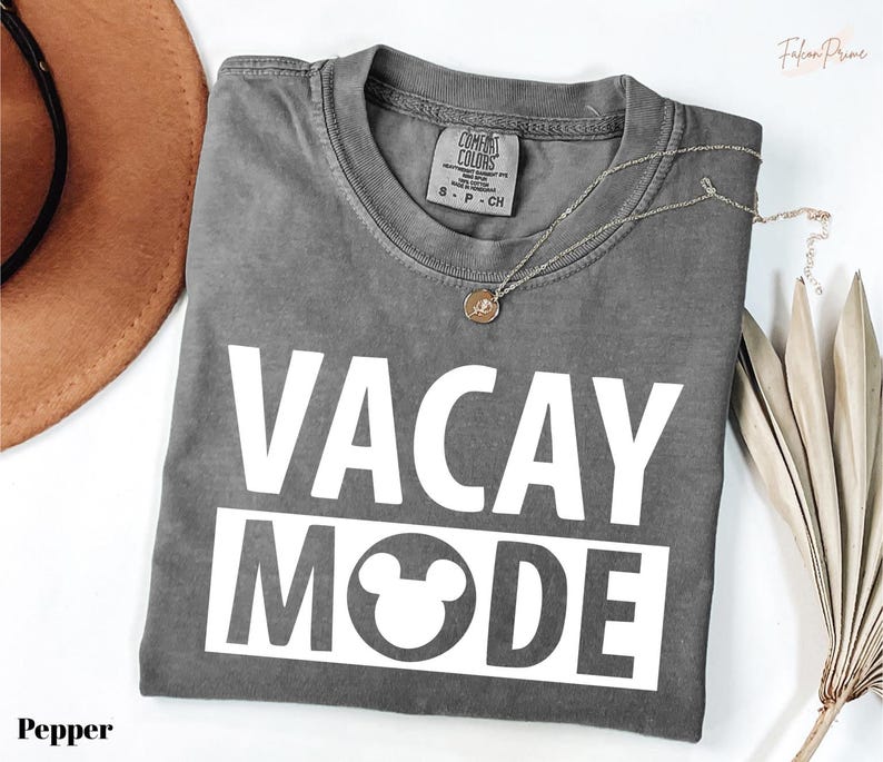 a gray t-shirt with the text "VACAY MODE" printed on it, along with a small pendant necklace. The t-shirt is laid out on a white surface, and there is a brown hat and some dried leaves nearby.