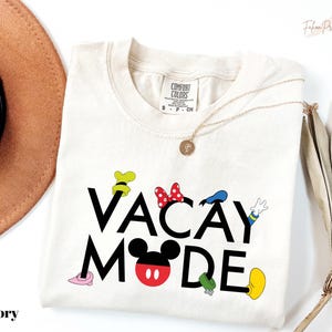 a white t-shirt with the text "VACAY MODE" printed on it, along with various Disney character illustrations. The t-shirt is laid out on a white background, and there is a brown hat and a fan visible in the background.