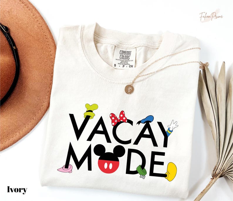 a white t-shirt with the text "VACAY MODE" printed on it, along with various Disney character illustrations. The t-shirt is laid out on a white background, and there is a brown hat and a fan visible in the background.