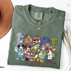 May include: Moss green t-shirt with a colorful graphic featuring Disney characters and the text "EPCOT Food & Wine". The design includes Mickey Mouse, Minnie Mouse, Donald Duck, Daisy Duck, and Goofy. The shirt has a crew neck and short sleeves.