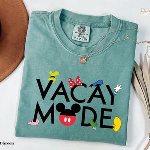 A light green t-shirt with the text "VACAY MODE" printed on it, along with various Disney character icons such as Minnie Mouse, Mickey Mouse, and Goofy. The t-shirt is laid out on a white surface, with a brown hat and some dried leaves visible in the background.