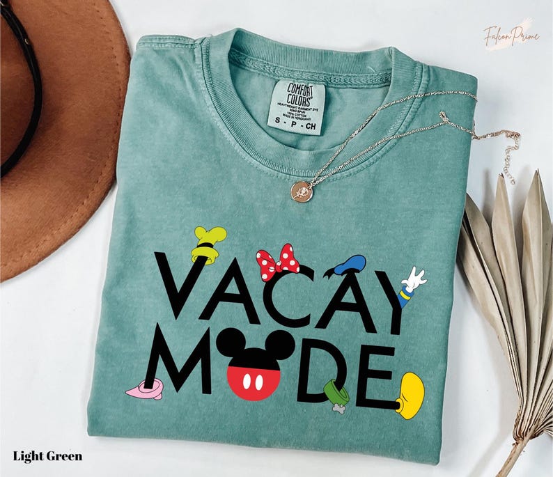 A light green t-shirt with the text "VACAY MODE" printed on it, along with various Disney character icons such as Minnie Mouse, Mickey Mouse, and Goofy. The t-shirt is laid out on a white surface, with a brown hat and some dried leaves visible in the background.