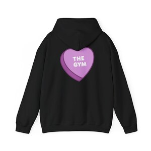 Puede incluir: Sudadera con capucha negra con un gráfico en forma de corazón morado en la espalda. El corazón presenta el texto "THE GYM" en blanco. La sudadera tiene capucha y mangas largas, adecuada para uso casual.